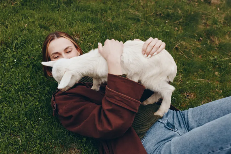 Relaxed woman lies on grass outdoors, embracing a white cat, enjoying a sunny day in casual clothing, a gentle, intimate lifestyle moment with affection and cuddling in a peaceful, candid outdoor