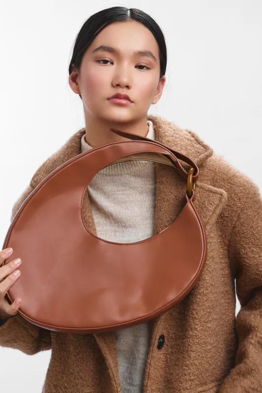 Asian woman wearing a cozy beige coat and knit sweater poses with a round brown leather bag, creating a chic studio portrait that emphasizes style and contemporary fashion.