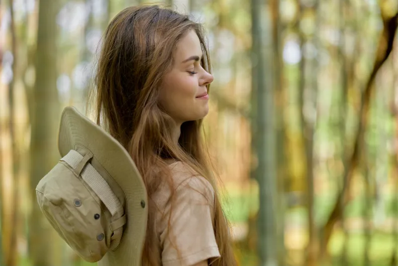Woman portrait in forest, smiling with eyes closed and hat draped on shoulder, relaxed outdoors scene. Young female with long hair enjoying peaceful nature moment in warm sunlight.