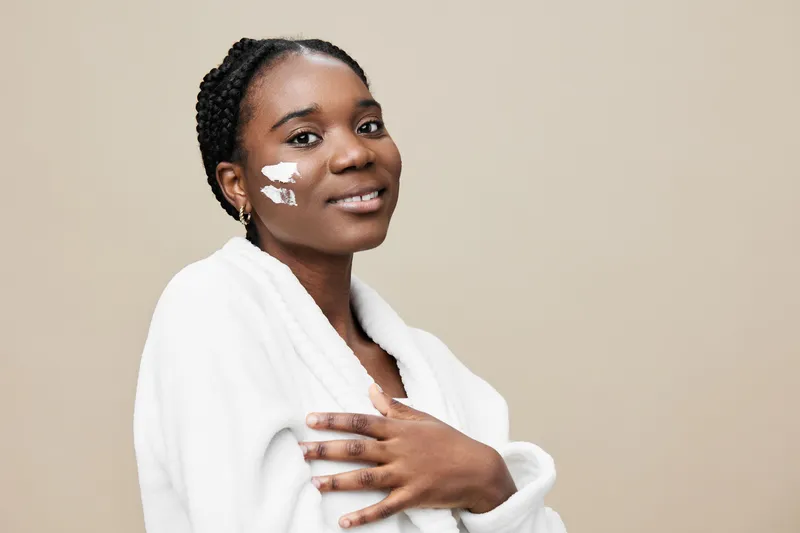 Beautiful African woman applying skincare cream with a smile, showcasing healthy skin in a minimalistic interior background