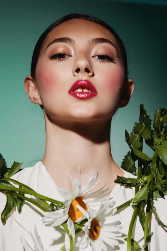 Young woman with bright red lipstick and dramatic pink blush wear a creative floral outfit made of daisies and greenery against gradient greenbackground light.