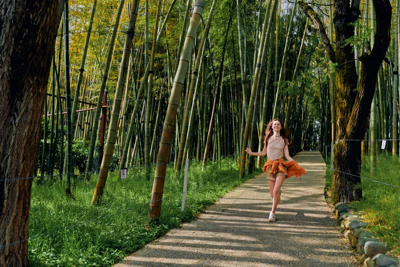 Woman girl bamboo forest path running in nature wearing an orange skirt, joyful motion and sunlight filtering through tall bamboo trees on a gravel trail for outdoor lifestyle.