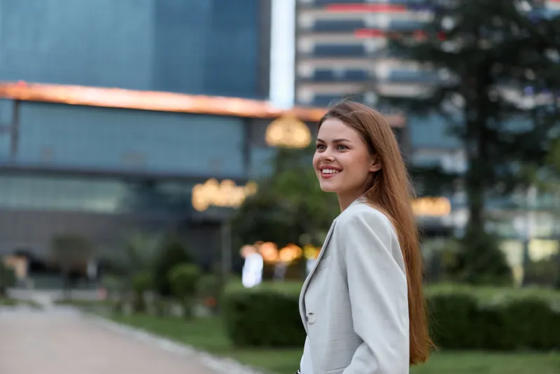 Confident business woman smiling outdoors, wearing a tailored suit, with a modern urban background and natural greenery, reflecting professionalism and empowerment.