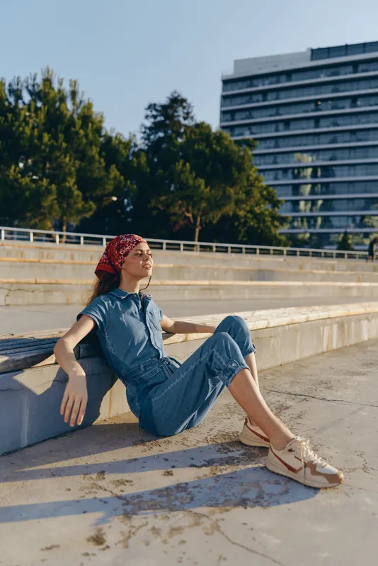 lifestyle woman in modern boho-western denim street style relaxing outdoors in film color, wearing red headscarf and casual sneakers on sunny day