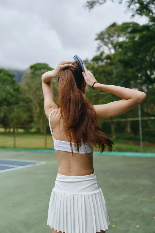 Young woman in sports attire brushing long hair outdoors in a green park, reflecting a healthy lifestyle and sporty vibe