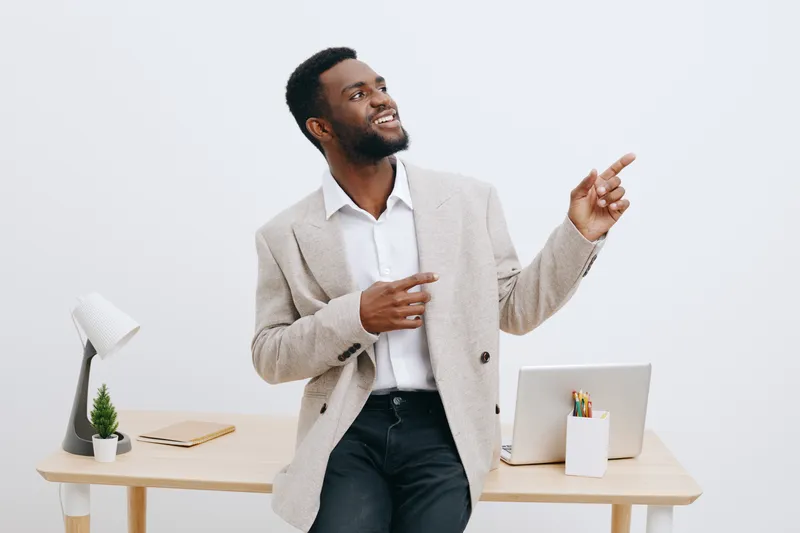 Full body smiling happy cheerful young African American man wearing beige blazer and white shirt sit at desk with laptop and stationery in modern office with plain light background studio shot. Busine
