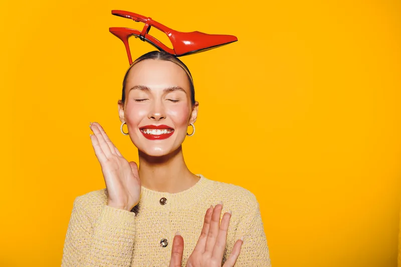 Smiling woman with red high heel shoes on her head poses with closed eyes and bright red lipstick on yellow background. Concept of fashion, fun, and playful mood in studio.