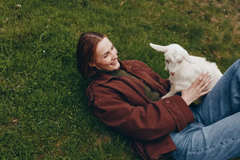 Young woman with long brown hair lies on grass smiling and gently holding a white baby goat enjoying a relaxed lifestyle moment outdoors in casual autumn clothing.
