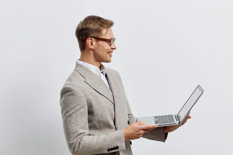 Smiling man wearing glasses and light gray blazer holding open laptop computer, looking at screen with smile, isolated on plain white background, studio shot, professional office attire, technology co
