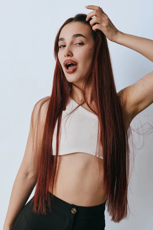 Young woman with long straight hair posing with hand in hair and mouth slightly open in casual white crop top and black pants against white background in studio.