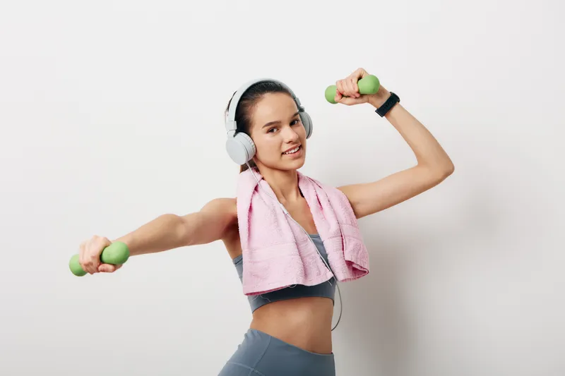 Fitness woman exercising with dumbbells, wearing headphones and a towel draped around neck, in a bright studio, energetic pose and athletic focus during a home gym routine.