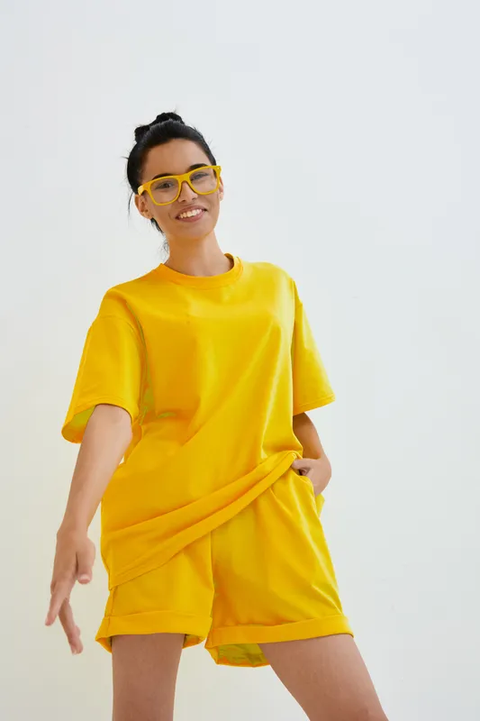 smiling woman in yellow casual outfit and glasses posing against plain white background, wearing shorts and t-shirt, youthful and cheerful mood