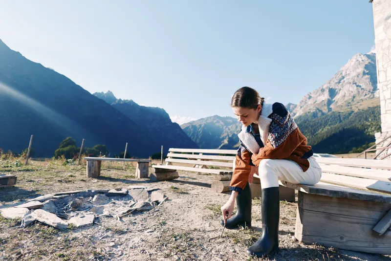 Woman sits on wooden bench in mountains wearing boots and warm sweater, holding a cup by a campfire site, enjoying nature and calm autumn outdoor camping moment at sunrise.