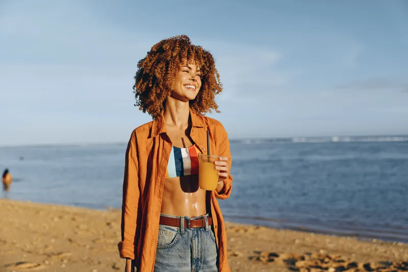 Woman with curly hair smiling on a beach by the ocean, wearing an orange jacket, denim shorts and casual style, outdoors in summer sunshine, relaxed travel lifestyle, soft natural light
