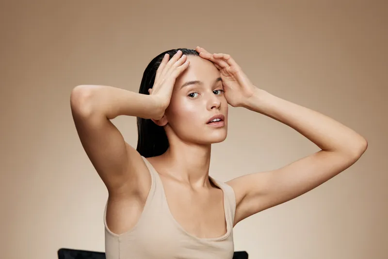 Young woman with smooth skin holding her head, showcasing healthy beauty and skincare, captured against a neutral beige background, emphasizing elegance and simplicity