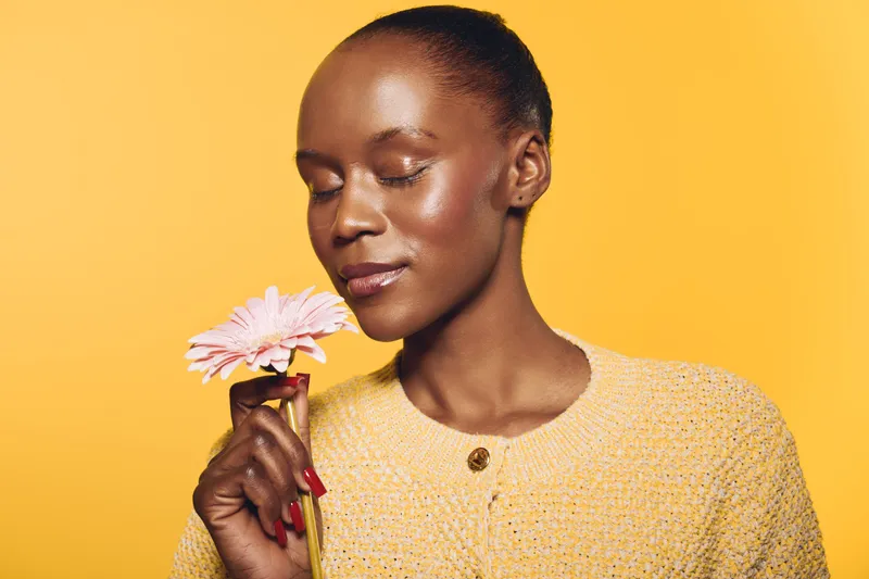 Close-up of a dark skinned African woman holding a pink flower near her face with closed eyes. She wears a yellow textured sweater, isolated on a bright yellow background. Concept of serenity.