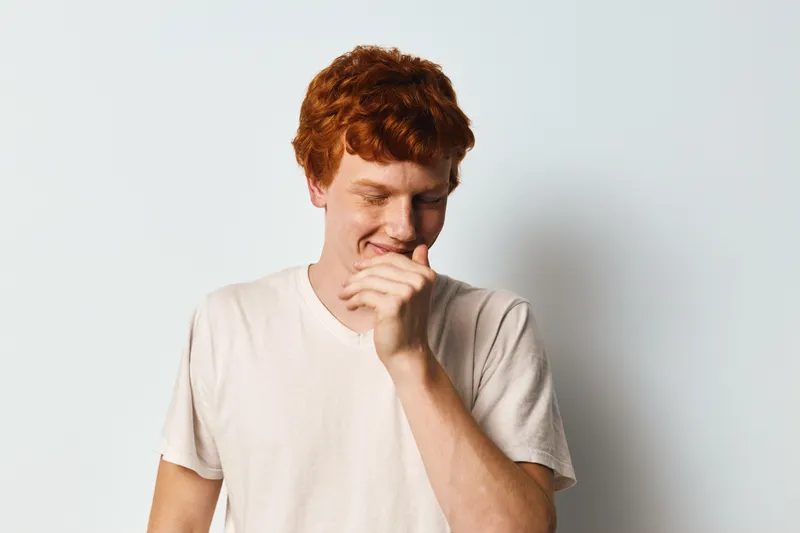 Young redhead man with curly hair wearing casual white t shirt standing smiling and looking down against plain white background in studio light.