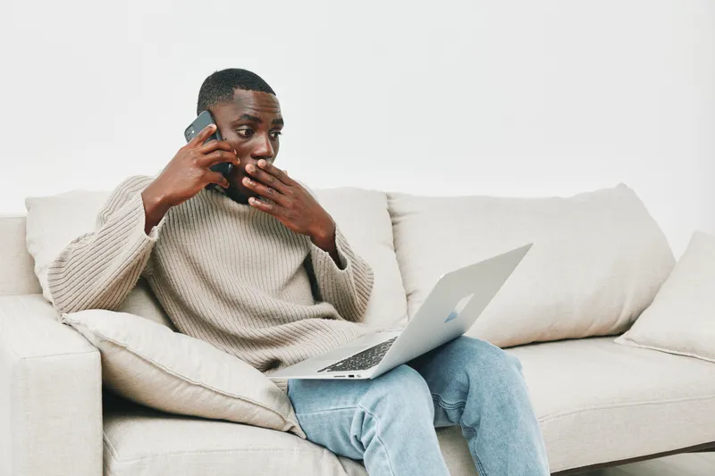 Young Black man in casual sweater talking on the phone while using a laptop at home, expressing concern and focus during a conversation