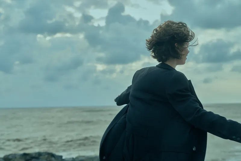 Business figure at the coast, coat fluttering in wind, facing the horizon over the sea, suggesting travel, contemplation, and open possibilities