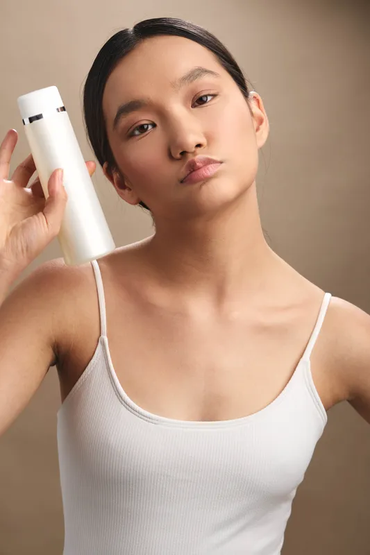 Asian woman in a white tank top holds a bottle near her face, posing in a minimal studio with soft lighting to convey clean beauty and calm confidence.