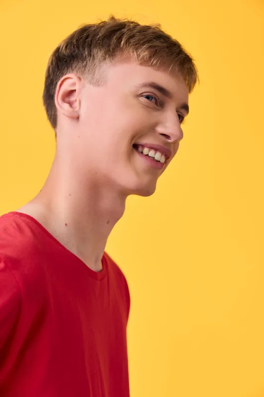 Smiling man in a red shirt against a vivid yellow background, side profile, casual fashion, cheerful mood, studio lighting, contemporary lifestyle photograph, bright and energetic vibe.