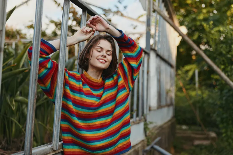 Woman in colorful sweater with smile, relaxed in greenhouse, portrait with closed eyes enjoying plants and natural light. Leisure, cozy knit, autumn mood and serene expression.
