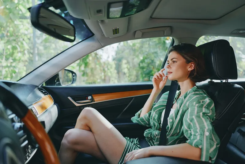 A stylish woman sits in a car wearing a seatbelt, gazing ahead with a calm expression as sunlight filters through the interior.