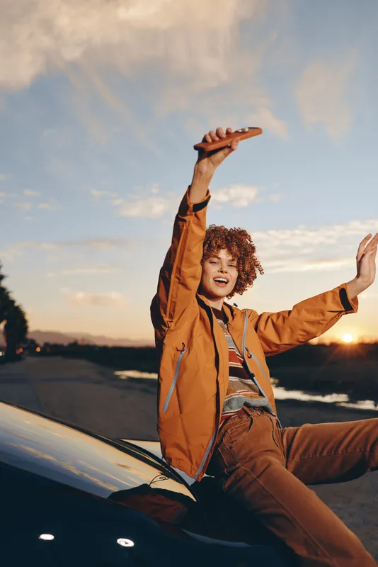 Cheerful woman lifestyle outdoors, sitting on car hood at sunset, orange jacket and curly hair, one hand raised, relaxed pose, golden sky, soft evening roadside background