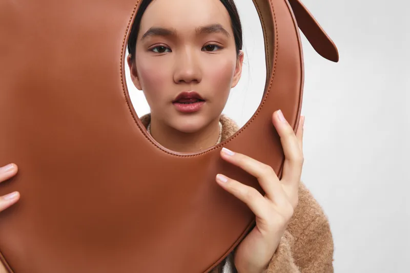 asian woman holds a circular brown leather bag close to her face in a studio portrait, soft lighting and a neutral backdrop create a chic fashion mood and poised expression.