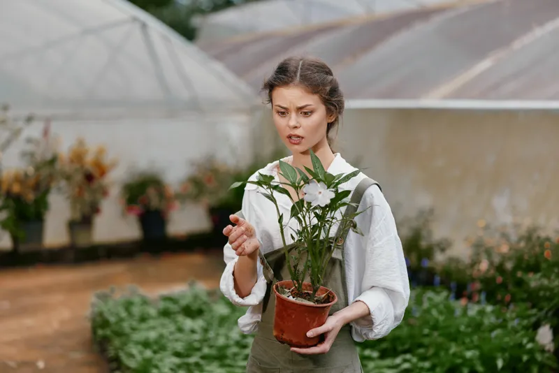 A young woman planting flowers in a greenhouse, showcasing a mix of emotions and passion for gardening while dressed in light, natural clothing