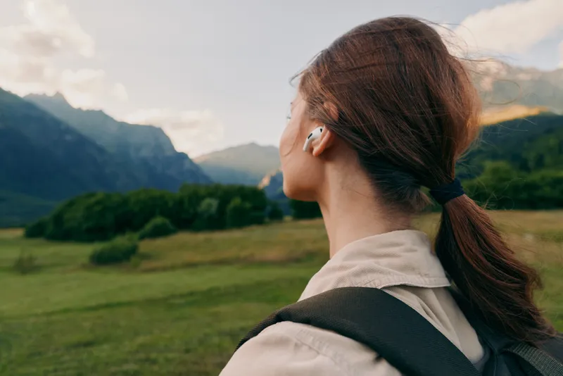 Woman with backpack stands in a mountain landscape, wearing headphones, gazing at distant peaks, enjoying outdoor life and serenity, travel inspiration for lifestyle themes