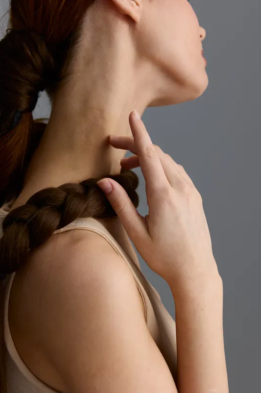 Close-up of woman’s neck and shoulder with delicate hand touch, showcasing smooth skin, braided hair, and soft neutral background in warm natural lighting.