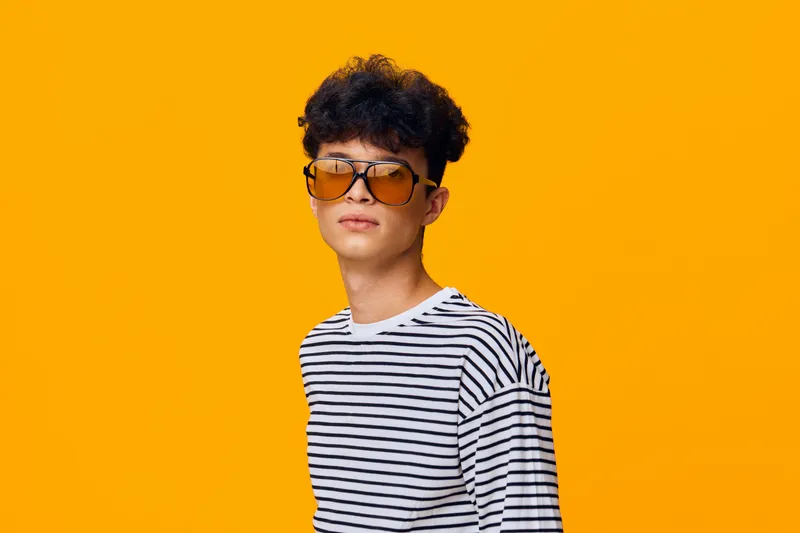 young man wearing striped shirt and oversized sunglasses posing confidently against vibrant orange background, casual style with curly hair and modern accessories