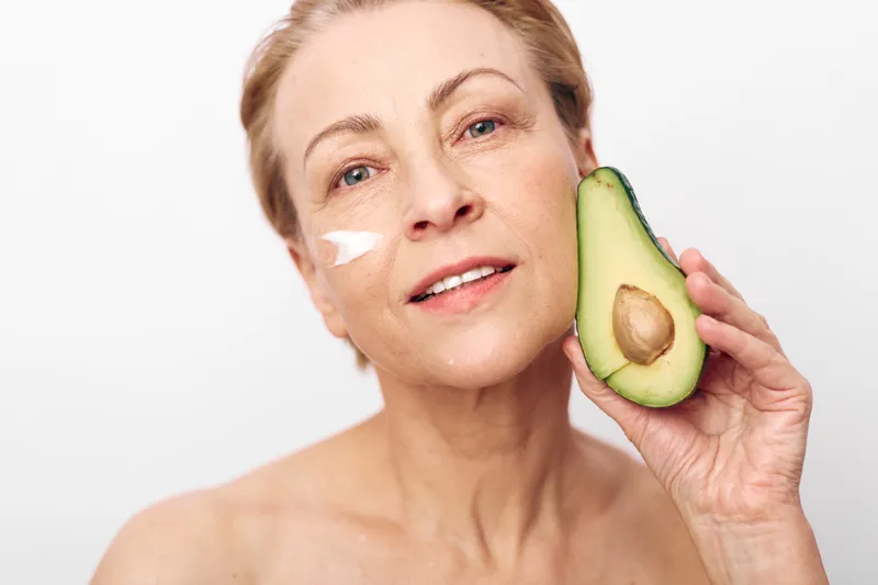 Mature woman with healthy skin holds fresh avocado near face with cream on cheek. Natural skincare and beauty concept focused on nutrition, wellness, and organic facial care routine.