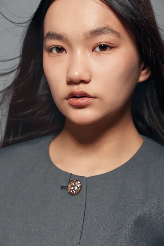 Portrait of young woman with natural makeup and smooth skin wearing grey top against plain background. Closeup of female face with soft expression and loose dark hair in studio light.