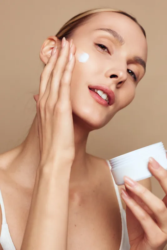 Woman applying face cream in closeup skincare moment, hands smoothing moisturizer on skin, beige studio background, soft light, beauty wellness hygiene concept