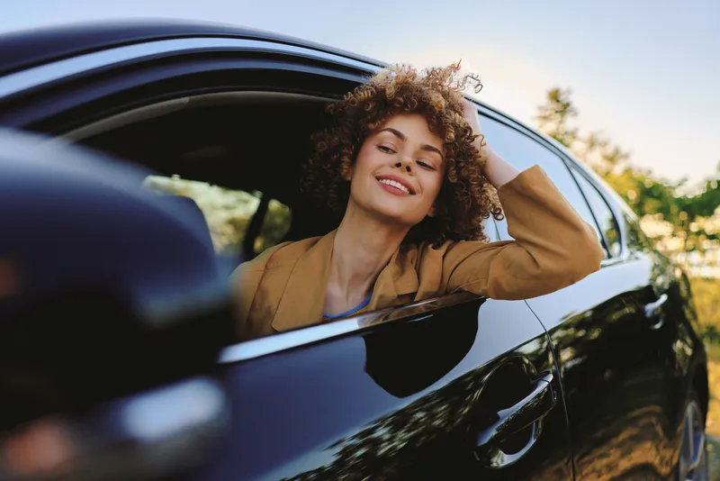Smiling woman with curly hair leaning out of a car window, retro film color vibe, casual lifestyle moment, warm light, candid travel mood, carefree expression, sunny day