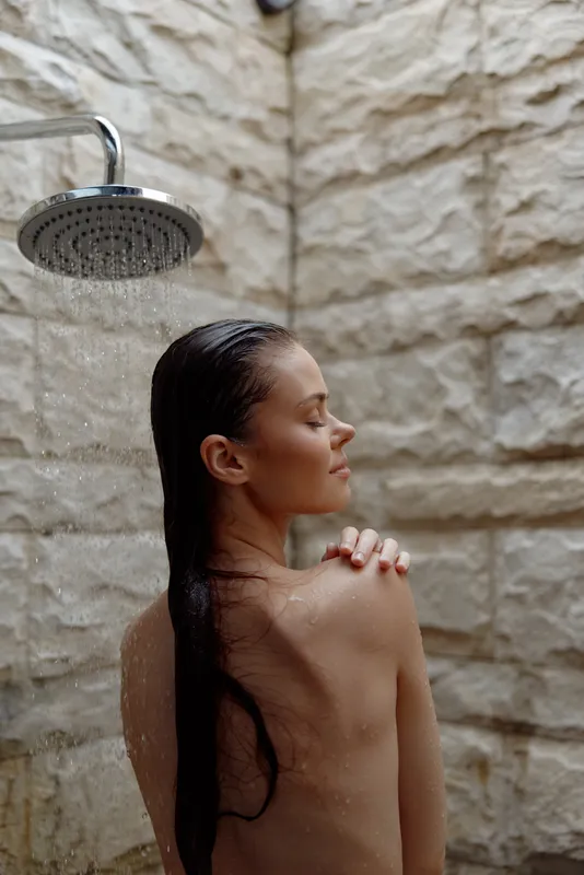 woman showering outdoors with wet hair standing under rainfall, serene expression, natural stone wall background, calm and refreshing moment
