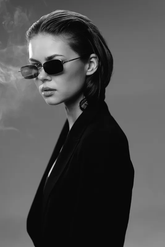 Black and white portrait of a serious woman wearing sunglasses and a dark coat with a smoky background.