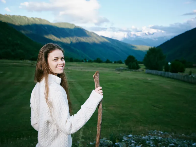 Woman holding a rustic stick in a green meadow with rolling hills and distant mountains, wearing a warm sweater, bright smile, outdoor scenery and tranquil countryside.