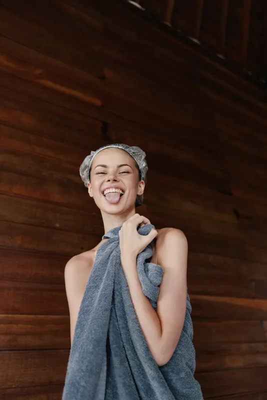 Playful woman wrapped in grey towel with hairnet making funny face against wooden wall, showcasing relaxed and joyful mood after spa treatment or shower.
