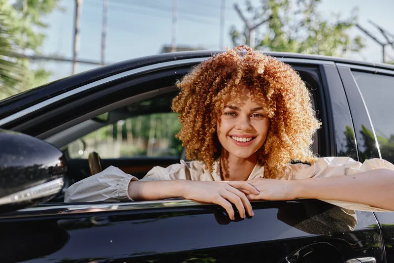Smiling woman with curly hair sitting in black car, enjoying sunny day outdoors, casual clothing, natural light, happy expression, summer vibe.