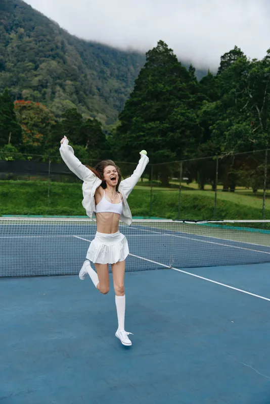 Happy young woman celebrating a tennis victory outdoors, wearing a stylish white outfit, with a scenic forest background and a joyful expression during the summer season