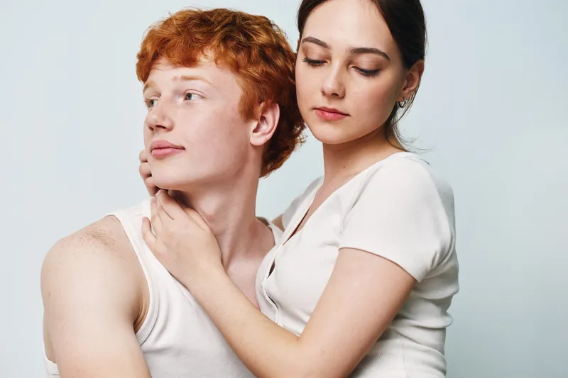 Couple, woman and man with red hair posing close, casual clothing with tender expression and gentle touch on light background. Young adults, intimacy and affection in studio.