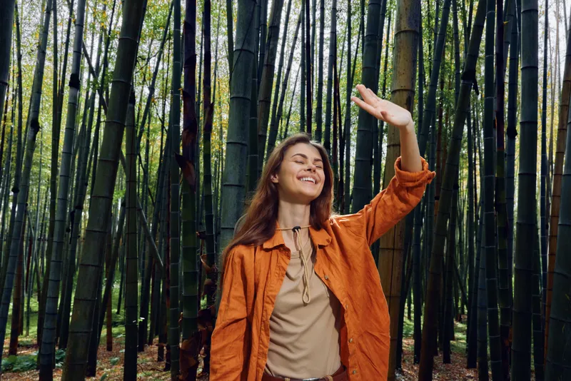Woman in orange jacket smiling in a dense bamboo forest, eyes closed and hand raised to warm sunlight among tall green stalks, enjoying nature, peace and a mindful outdoor moment.