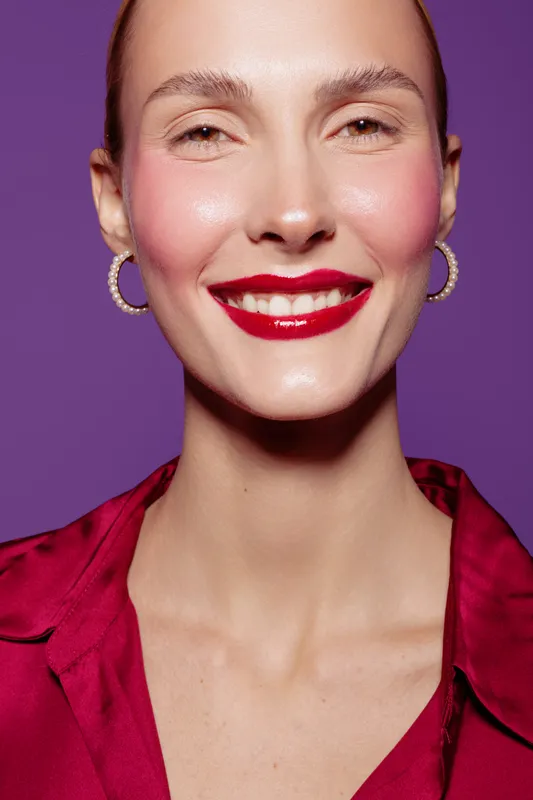 Smiling woman with red lipstick and hoop earrings poses against purple background. Close-up portrait with smooth skin and bright makeup. Concept of beauty, fashion, and confidence.