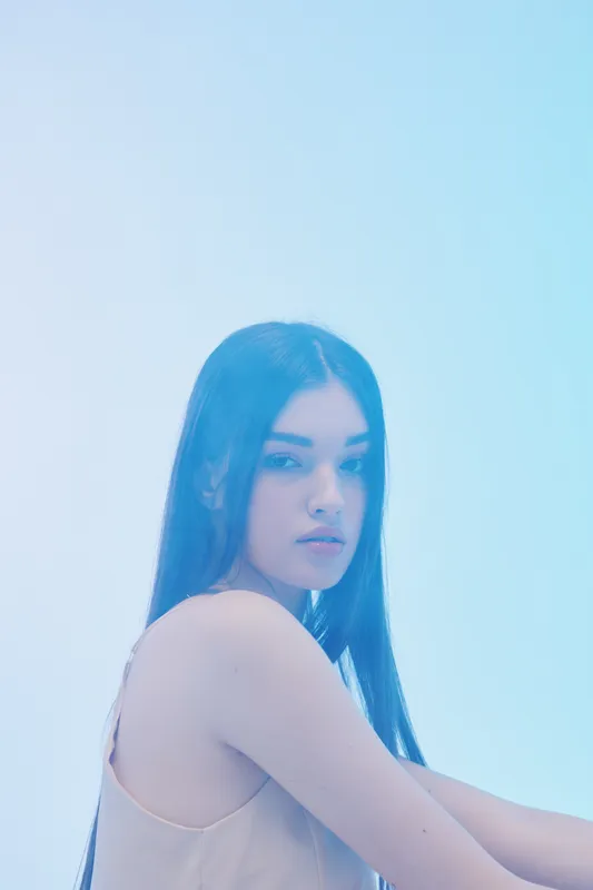 Young woman with long dark hair poses in soft lighting against a pale blue background. Concept of beauty, fashion, elegance, and modern style with minimalistic mood.