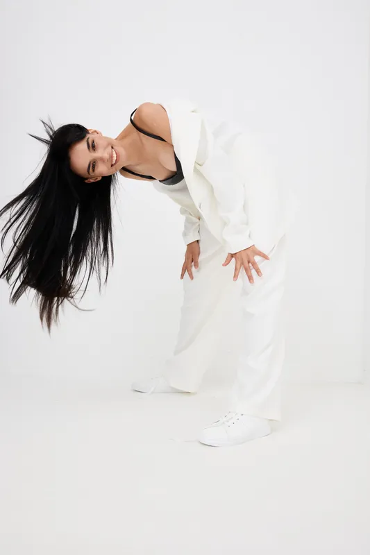 smiling woman in white suit bending forward with long black hair, casual and playful pose, studio background, bright stylish outfit, joyful energy