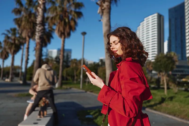 Young woman with curly hair wears a red jacket while using her smartphone outdoors on a sunny day near palm trees and modern buildings.