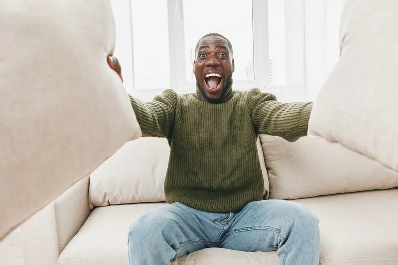 Happy African American man in cozy sweater cheerfully embracing pillows on a light couch, showcasing joy and comfort in a bright living room
